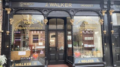 The outside of John Walker's shop in Stockton. Signs in the left window read 'Quinn's nerve and brain tonic' and ' Beecham's cough pills'. On the right gold letters say 'Dr Brookes' cod liver oil' and 'soothing powders'.