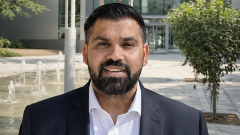 Mann is stood outside a building with a water fountain and small tree behind him. He has a navy suit on and a white shirt. He has dark hair and a beard.