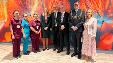 The image shows eight people, five women and three men standing in a curved line smiling for a photograph. Behind them is a colourful wall with painted vibrant flowers.