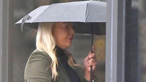 A woman with long blonde hair and wearing makeup holds a black umbrella over her head as she walks into a building. She is wearing a green coat and black top.