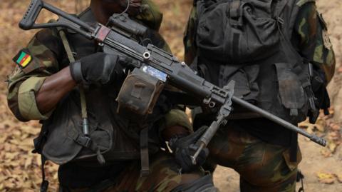 A man in army fatigues holds a gun. He wears a badge bearing the Cameroonian flag.