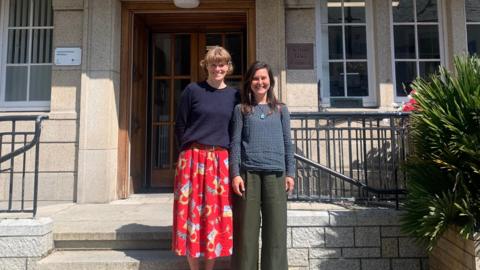 Laura Peach and Rosana Cox stand on the stone steps of the St Ives Guildhall smiling at the camera in the sunshine