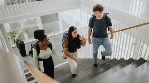 Three students wearing rucksacks on their backs chatting happily with each other as they climb steps inside a building.