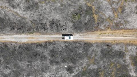 A white pick-up truck drives along a gravel track surrounded by blackened heath following the severe fire 12 months ago