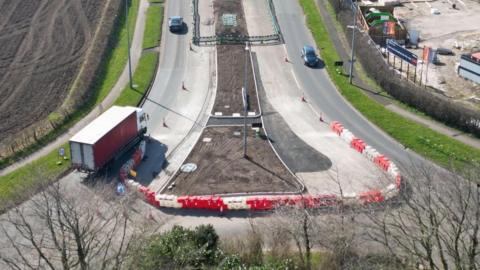 Roadworks on a dual carriageway showing red and white barrier sectioning off a new layout. Two cars and a lorry are travelling along the road.