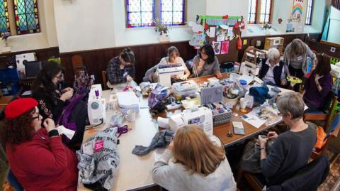 Ten people sat around a table using sewing machines to repair clothes. They are in a church hall with dark wood panels half way up the walls. There are stained glass windows in the wall