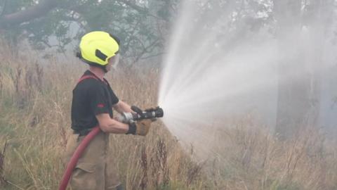 A male firefighter wearing a yellow helmet, a black T shirt and beige trousers holds a large red hose and sprays water at moorland grass