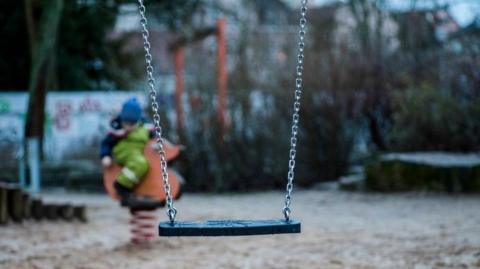 A generic image showing a child's swing in the foreground. A child, who is blurred on the photo and wearing a blue hat, is in the background sitting down. 