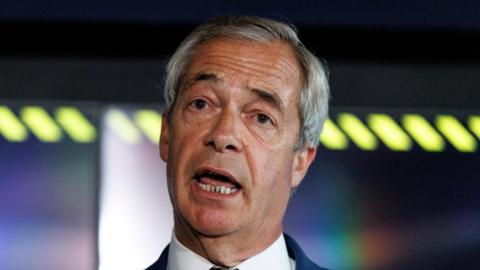 Nigel Farage, wearing a white shirt and navy blazer, tilts his head to one side as he speaks