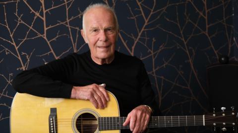 Allan Taylor seated and looking at the camera and smiling with a guitar o his lap and both arms resting on it.