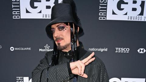 Boy George is wearing a pinstripe jacket and a black bucket-style hat with tassles. He has facial hair, colourful eye make-up gems around his eyes. Behind him is a black wall with the logo 'British LGBT Awards' on it. He is making a peace sign with two fingers.