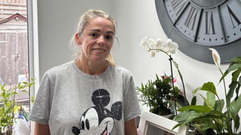 The image shows a woman with blonde hair, wearing a grey t-shirt. She is standing indoors in front of pot plants. There is a big clock on the wall.