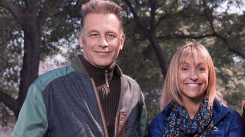 The Winterwatch presenters Chris Packham and Michaela Strachan are standing side by side in a professional picture. Mr Packham has short brown hair and is wearing a brown zip-up fleece, and a blue/ green jacket. Ms Strachan has short blonde hair with a fringe and is wearing a dark blue coat with a patterned scarf. 