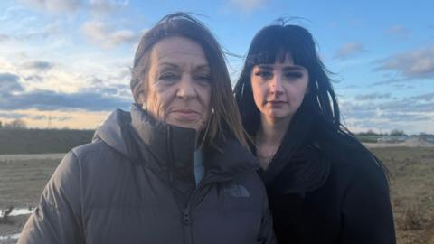 Jessica and Nicole Groucott are standing next to each other, Nicole on the left is wearing a black zipped up coat and she has medium length brown hair. Jessica, who is next to her, has long black hair and is wearing a black coat. The pair are in a field and the skies behind them are filled with clouds while sunset descends. 