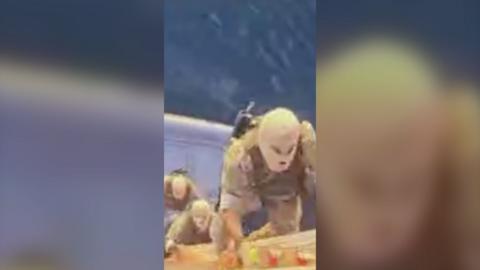 Masked men in beige fatigues climb up a ladder on the side of a ship.