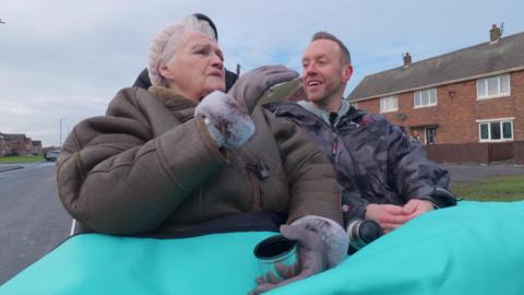 A woman with grey hair wearing a brown coat sit on a trishaw next to a man with short brown hair, wearing a blue jacket.