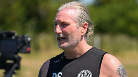 Forest Green Rovers boss Robbie Savage wearing a black vest with his initials on, and the club logo on, with his left arm covered in the sun, revealing his tattoos. 