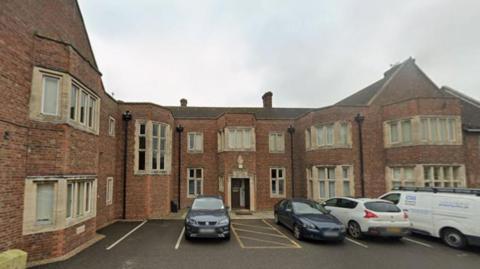 The exterior of the Health Authority Offices - a large red brick building with multiple stone window casings. There are three cars - one grey, one blue and one white - and a white van parked in front of the building. The sky is overcast.