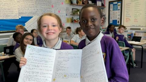 Maxwell has blonde hair, tied back, while Rayleigh has short black hair. They are both wearing white polo shirts and purple jerseys, embroidered with the school logo. They are holding their A4-sized workbooks open. Other children are sitting at desks behind them. School work is displayed on the classroom's walls.