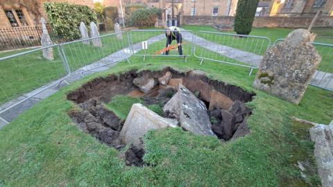 A stone box tomb which has collapsed into a large hole in the ground, through the soil and into the crypt below, a small stone home buried underground. There are metal barriers around the hole and a man in a high-vis jacket. There are other gravestones very close to the edge of the hole, which is about 12ft (3.6m) wide.
