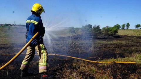 A firefighter in a navy blue top with a yellow hi-vis strip and brown trousers with the same yellow strips is standing in a field. They are wearing a yellow helmet and holding a yellow hose which is spraying water over blackened grass.