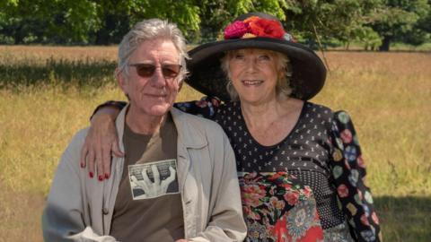 Gavin Petrie and Jan Etherington in a field. He is on the right and wearing sunglasses and a brown top and cream overshirt. Jan has her arm around him and is wearing a big black hat with flowers around the brim 