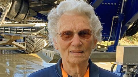 Shelia Warner stands in front of the base of a lifeboat. She has curled white hair and wears tinted glasses. 