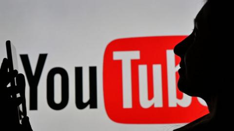 Woman holding a smartphone in front of a YouTube logo displayed on a computer screen