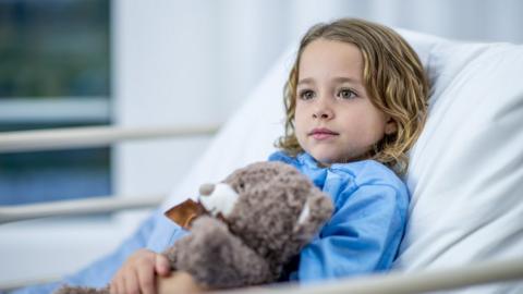 A girl lies in a hospital bed and looks across the room with a noble, innocent expression. She is holding a teddy bear in her arms and wearing blue hospital clothing.