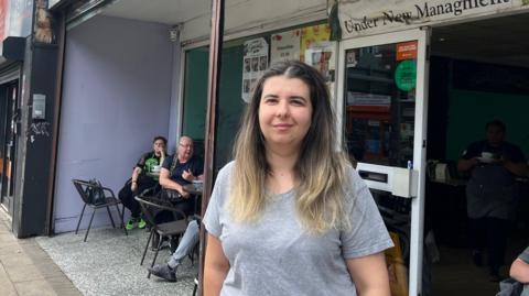 Niya Konakchieva, who is smiling, wearing a grey t-shirt and has long brown hair with blonde highlights, stands outside a cafe where customers can be seen sitting on outdoor chairs. 