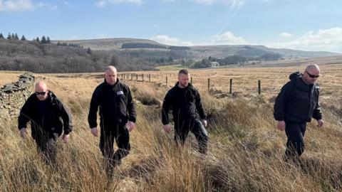 Four men dressed all in black walk through long, dry grass in between a wire fence and stone wall through a sloped field. Moorland and forest can be seen in the distance on a bright, sunny day. 