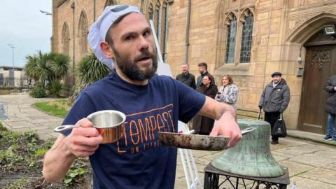 Lee Johnson had brown hair and a brown beard. He is wearing a chef hat and a blue t-shirt which says Tempest on Tithebarn in orange writing. He is holding a frying pan and a silver pan.