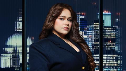 A woman wearing a dark coloured blazer. She is stood in front of a set of glass windows, with a city skyline visible behind her.