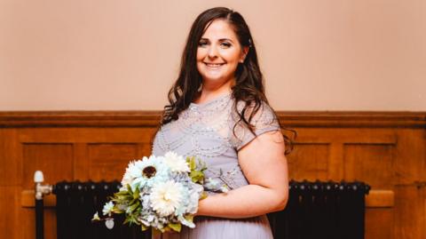 A smiling Laura-Jane Seaman looks directly at the camera, she is holding a bouquet of flowers and wearing a pale grey dress with beaded embroidery. 