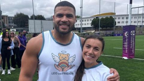 Zack George wearing a white vest with a motif of a yellow teddy bear with wings and a halo, and a pair of hands opening to reveal a rainbow. He is standing with his arm around his wife Samantha who is wearing a white t-shirt. They are both smiling. 
