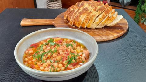 Jack Stein's hake and chickpea stew. In the foreground, a bowl of orange and red stew with green parsley garnish. In the background, a sliced loaf of crusty bread on a wooden board.