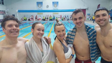 A group of swimmers stood in front of a pool. Three men can be seen in swimming trunks and two women with towels on. They are all smiling into the camera.