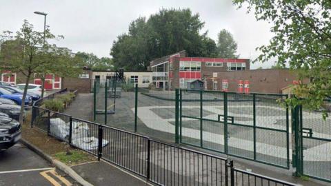 The outside of Red Hall Primary School in Darlington. There is a high green metal fence marking the boundary and the school is made of bricks with red panelling around the windows. It is a mix of late 20th Century single and two0-storey buildings with flat roofs, all interlinked. A central entrance painted a cream colour sits between two of the buildings. There is a car park to the left, outside the green fence, and several vehicles parked there are partially visible.