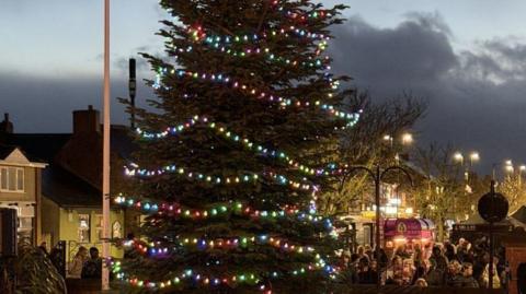 A large Christmas tree has layers of multi-coloured lights all over it. A Christmas market is packed with people between different food stalls in the town square. The sky is grey and cloudy behind it.