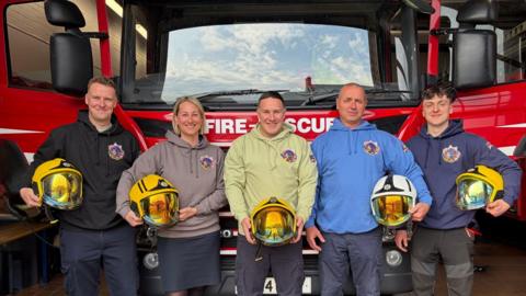 Five firefighters - including one woman and four men - stand in front of a fire engine while holding helments.