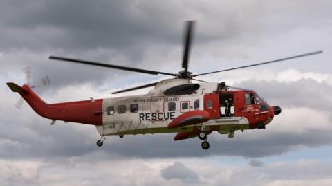 A red and white helicoper in the sky with the words "Irish Coast Guard Rescue" on the side.