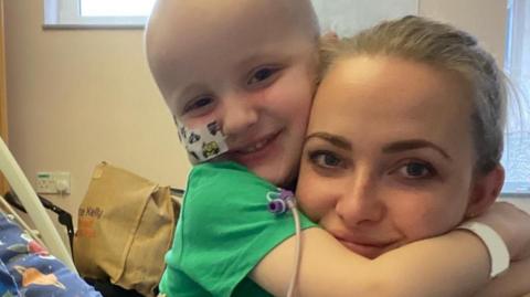 A young boy with no hair and a tube going into his nose, hugging his mum. They are both smiling.