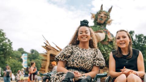 Two women sit in chairs in a field, smiling at the camera. Behind them is a big green man sculpture