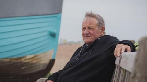 A man wearing a black hooded top and grey shorts. He is sat on a bench with a blue boat on a pebbled beach behind him.