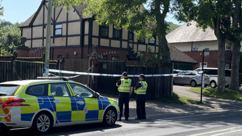 A police car and two police officers are stood outside crime tape which is in front of a large gate, in a residential area. 