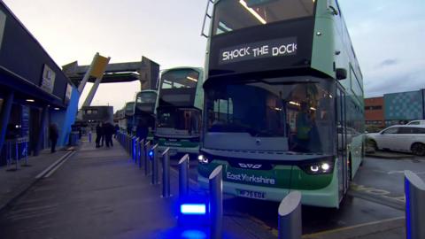 Several green buses are lined up outside The Deep aquarium, one of them is displaying 'shock the dock' on its display board.