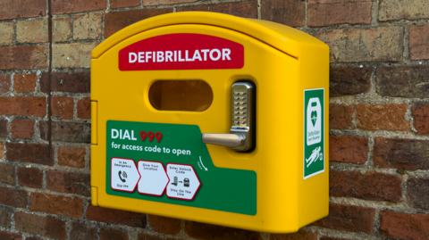 A yellow defribullator box mounted on a brick wall with the word "Defibrillator" in red on it, along with instruction diagrams on its use.
