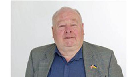John Allen. He is wearing a grey suit with a blue shirt. He has a round face and grey hair, which has receded on top. He is an older man.