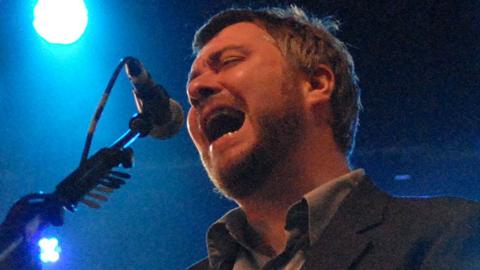 Singer Jimi Goodwin, with short hair and a beard and wearing black suit jacket and grey shirt, sings into a microphone in front of bright blue lights