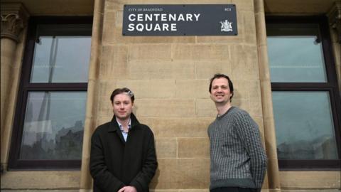 Two men stand below a smart black sign saying City of Bradford Centenary Square, which also features Bradford's civic crest